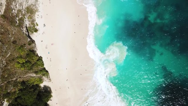 Drone Shot directly above KelingKing Beach on the island of Nusa, Penida, Indonesia. Tourists can barely be seen on the beach while waves of crystal clear blue water crash on the shore.