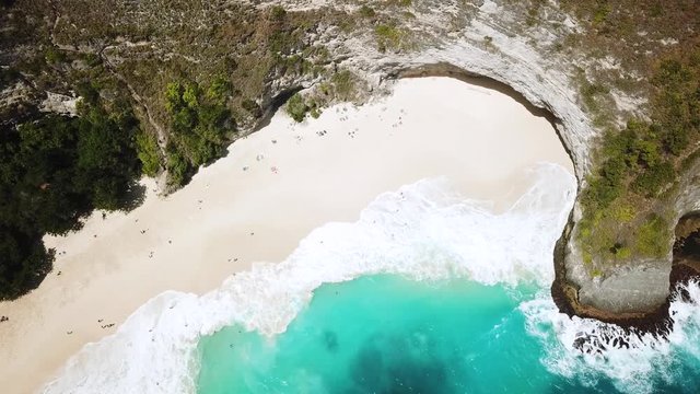 Bird's Eye drone shot above tourists relaxing on KelingKing beach while large waves crash onto the shore. This is off the coast of Nusa Penida, Indonesia.