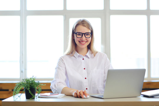 Young blonde beautiful girl in glasses and white shirt working on laptop in office