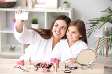 Mother and her little daughter in bathrobes taking selfie while applying makeup at home