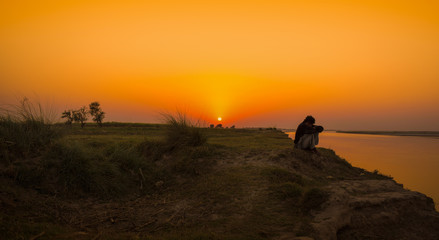 landscape image of a man sitting on a river bank at sunset time