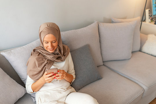 Elegant attractive female muslim using mobile smartphone searching online information when she sitting on sofa couch relaxing. Portrait of happy woman using mobile phone while sitting on a couch