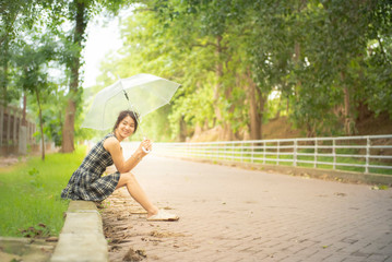 Happy asian woman teenage in casual dress sit by walkway with big smile hand hold umbella in public park and copy space.
