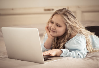 Beautiful cute little blonde girl playing and surfing the internet on laptop at home.