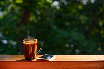 Cup of coffee and cellphone on the windowsill over blurred forest background at sunny summer day. Free copy space.