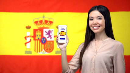 Female holding cellphone with learn Spanish app, flag on background, education