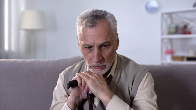 Old disabled man with walking stick sitting on couch, diseases at retirement age