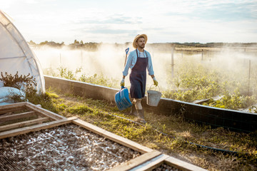 Well-dressed farmer with working tools standing on a farmland with automatic watering on a farm during the sunset