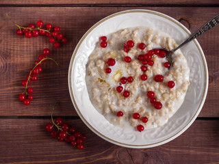 Oatmeal on milk with red currant in a white plate on a wooden brown tray, a scattering of currants on a tray, a tablespoon in a plate