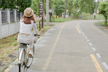 Pretty girl with straw hat is happy riding with bike down wide beautiful park alley with trees around on sunny summer day.