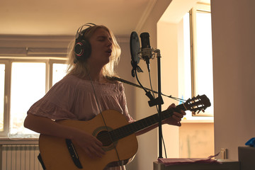 Beautiful blonde singer girl in headphones with a guitar in home recording studio sings a song into...