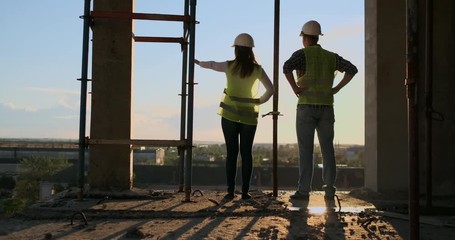 Back and side view of caucasian builder in White helmet and green vest looking drawing standing on unfinished construction background