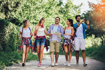 Obraz premium Group of smiling friends walking with backpacks in woods - adventure, travel, tourism, hike and people concept