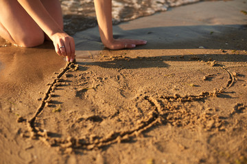 The girl is drawing a heart on the sand by the sea in the evening with fingers