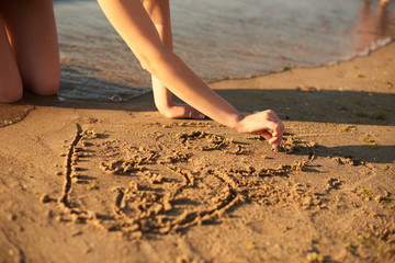 Fototapeta na wymiar The girl is drawing a heart on the sand by the sea in the evening with fingers