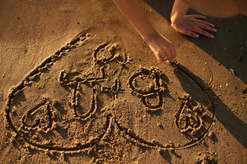 The girl is drawing a heart on the sand by the sea in the evening with fingers