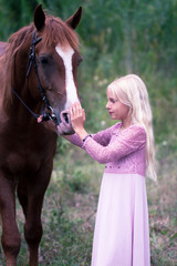 innocent blonde girl looking at the horse in forest. beautiful Caucasian girl with long blonde hair in a pink dress hugs a brown horse. innocent childhood concept. natural beauty. hipster style.