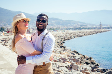 Fototapeta na wymiar smiling happy european woman in straw hat and sexy pink dress walking with afro american ethnic man by summer beach with rock view in Turkey tropical resort