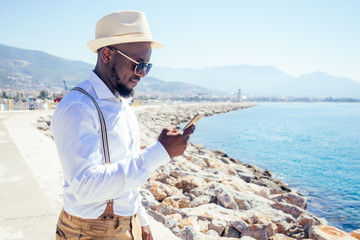 Fototapeta na wymiar handsome african american person casual fashion look outfit in straw hat,white shirt walking by Tyrkey coast and using modern smartphone route gps navigator map application