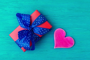 Red gift box with wide blue spotted ribbon and pink heart on turquois wooden background