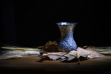 Still life against dark background. Metal vase standing on dry leaves and stalks of wheat