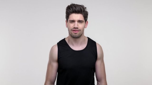 Serious young bearded sporty man in black shirt disagree with someone, shaking his head and gesturing with hands while looking at the camera over gray background isolated