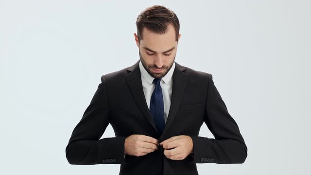 Serious young businessman in black suit and blue tie making few steps and buttoning his jacket while looking at the camera over gray background isolated
