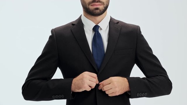 Cropped view of serious young businessman in black suit and blue tie buttoning his jacket and putting his hands in pockets over gray background isolated