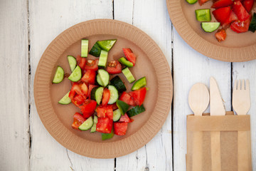 paper cardboard dishes with vegetable salad tomatoes cucumbers with wooden devices fork horse knife on a white wooden table