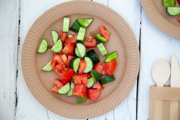 paper cardboard dishes with vegetable salad tomatoes cucumbers with wooden devices fork horse knife on a white wooden table