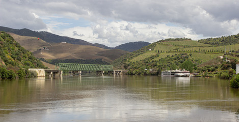 Landscape of the Douro river in Portugal. You can see a cruise ship docked next to an old railway bridge and numerous fields of vines. - obrazy, fototapety, plakaty