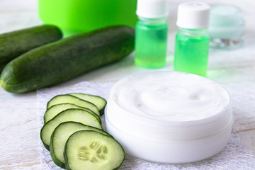 Face cream in a white jar with cucumber extract next to slices of fresh cucumbers on a white wooden background.