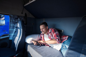 Truck driver lying on bed in his cabin communicating with his family via tablet computer. People...