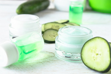 Face cream in a glass jar with cucumber extract next to slices of fresh cucumbers on a white wooden background.