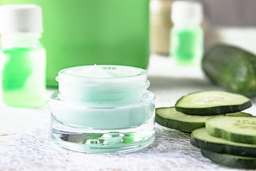 Face cream in a glass jar next to slices of fresh cucumbers on a white wooden background.