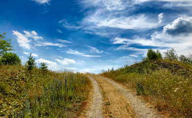 Rural dirtroad going uphill surrounded by trees and grass fields under a clear blue sky with white fluffy clouds