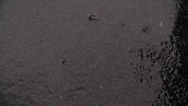 A slow motion shot of rain falling from the sky onto a dark grey rough concrete floor, the raindrops splatter with every drip. The floor is already soaked from the torrential downpour. 