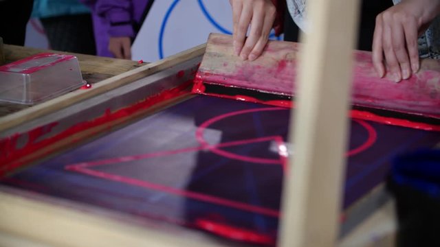A close of shot of a screen printing frame, a child is being shown how to use this printing technique by an artist. The red paint gets pushed over the screen to create the print below
