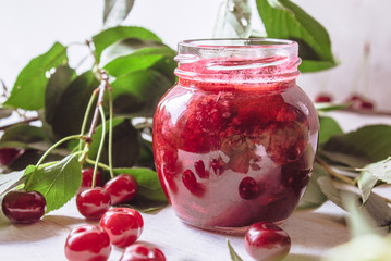 Bank with homemade cherry jam on a wooden background near the berries and leaves.