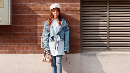 Street photography of a cute Chinese young woman in jeans and white hat with red brick wall background in sunny day, female portrait.