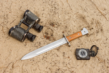 Knife bayonet with wooden handle lies on the sand with old military binoculars and old compass....