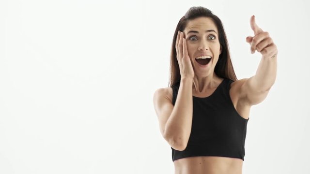 Cheerful beautiful sporty brunette woman in black sportswear becoming surprised, touching her face with hands and pointing on something with finger over white background isolated
