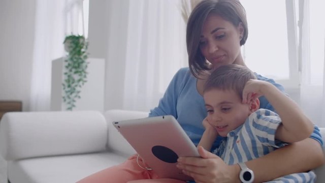 Mother and son sitting on sofa using digital tablet. Happy mom and little boy using tablet with touchscreen together watching a video. Smiling mother and cute boy playing on digital tablet