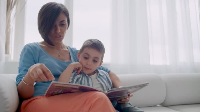 Happy mother and child son reading book laughing in bed. Happy family mother and child son reading holding book lying in bed, smiling mom baby sitter.