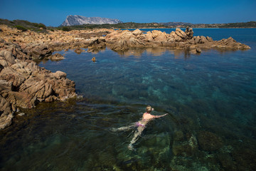 Aerial view of swimming woman in the sea. Mediterranean sea in Sardinia, Italy. Summer seascape with girl, clear azure water, waves at sunrise. Transparent water. Top view with cliffs and mountains