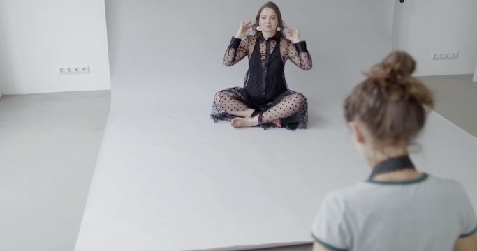 Backstage of the photo shoot: Beautiful caucasian pregnant model sits on studio floor posing and straighten her hair  in a Moment photographer starts taking photos.  Studio photoshoot with professiona