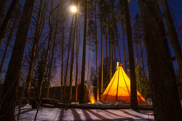 Teepee in the forest at night. A lighted teepee in a dark forest. National Indian housing. Winter overnight in the forest. Fire near the home of the Indians. Yurt, trees and moon.The light in the tent © Grispb