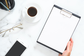 Modern home office desk workspace with blank paper clipboard in woman hand, coffee cup, glasses, smartphone, stationery on white marble background. Flatlay, top view.