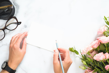 Woman hands holding pencil and write on blank paper card mockup over modern home office desk workspace with stationery, rose flowers bouquet, glasses, Flat lay, top view.