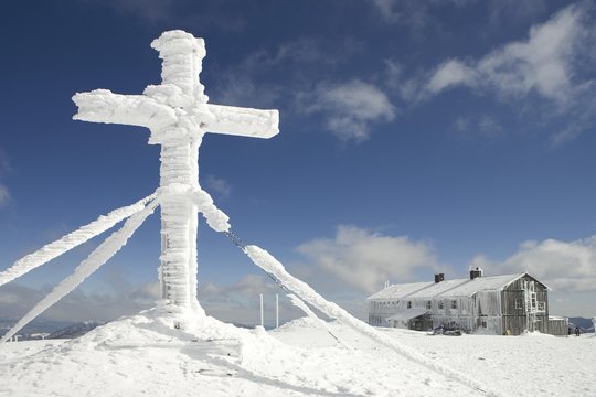 Snow covered cross and the Alois G&cedil;nther mountain hut on top of the mountain Stuhleck Styria Austria